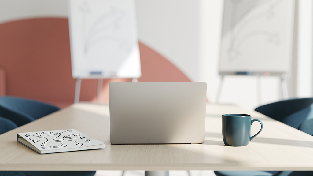 A sunlit meeting table with an open laptop, a notebook with flowing arrow diagrams and a petrol-blue mug, a curved coral